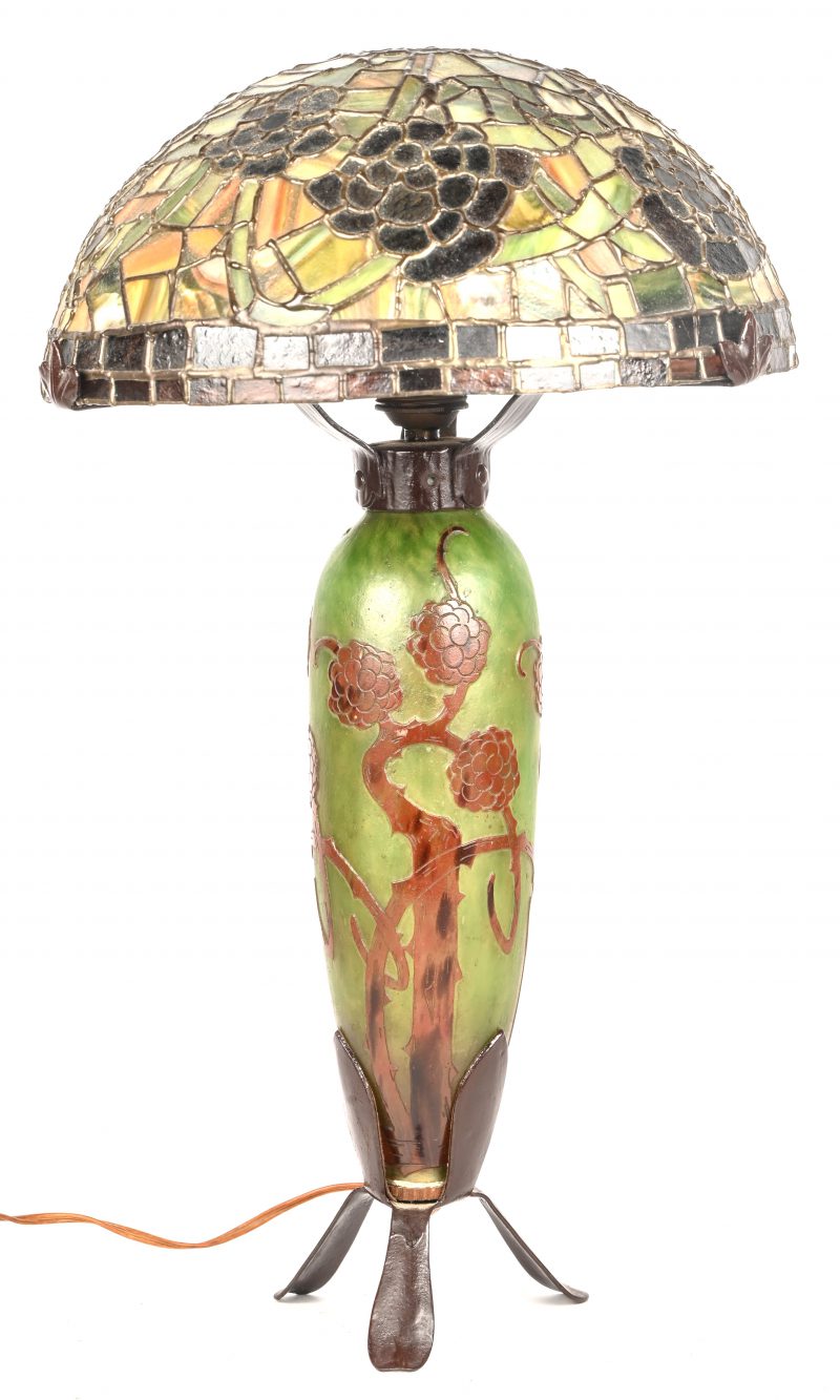 Een tafellamp met een glas-in lood kap in de geest van Tiffany’s. Lampenvoet van glaspasta versierd met distels. Klein letsel aan de rand van de kap, door een beugel.  Medio XXste eeuw.