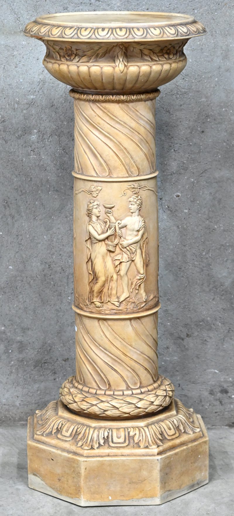 Een zuil van monochroom beige aardewerk met een reliëfdecor van personages naar de Griekse oudheid.