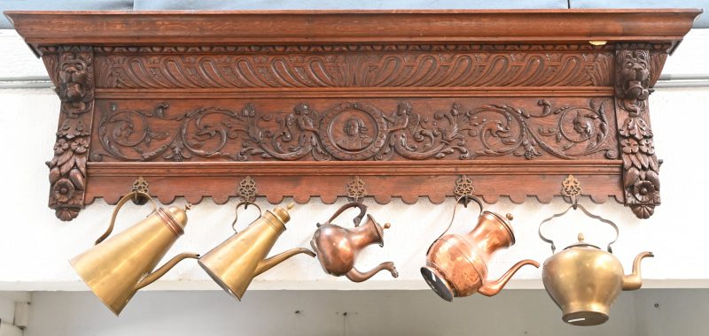 Een gebeeldhouwde eikenhouten kapstok versierd met leeuwenkoppen, met vijf koperen haakjes, waaraan vijf diverse koperen waterketels en koffiepotten. Omstreek 1900.