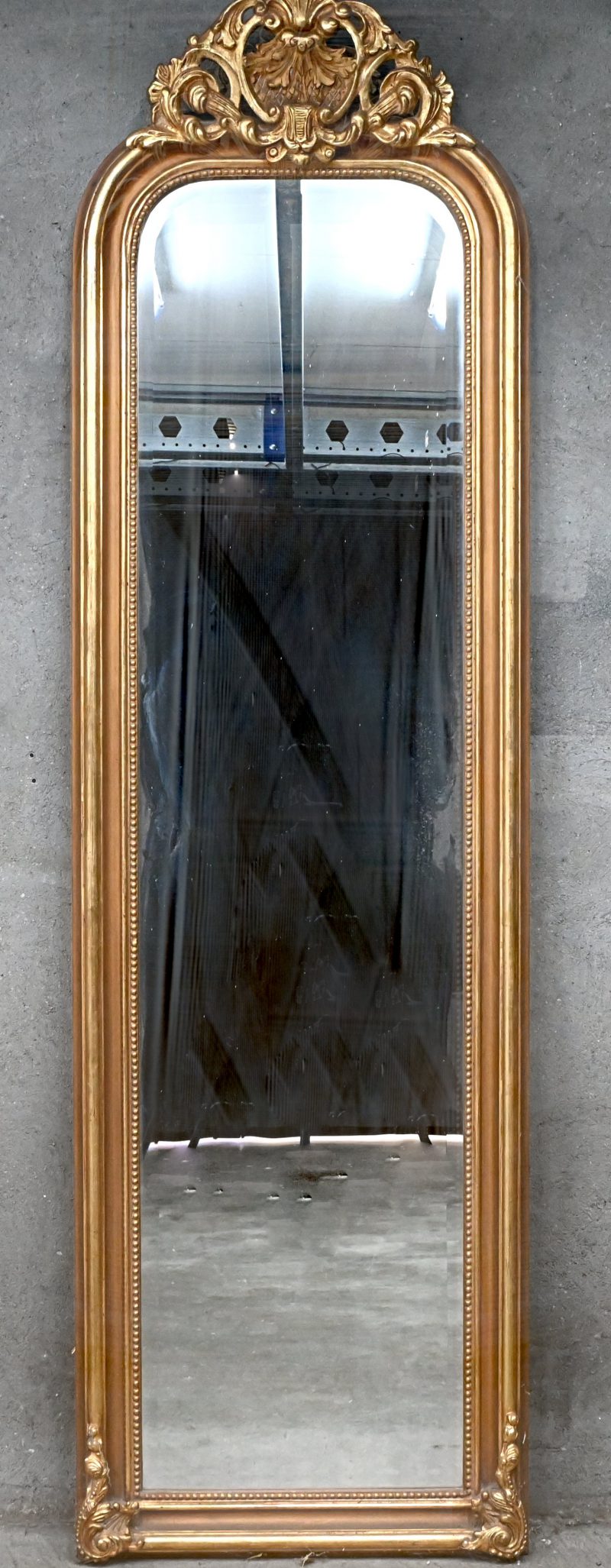 Een geslepen manshoge spiegel in goudkleurige kader, met ornamenten.