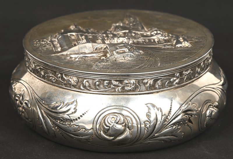 Een ronde doos van gedreven zilver. Op het deksel een stadszicht, op de buik bloemen en guirlandes. Nederlandse keur, tweede gehalte (835).