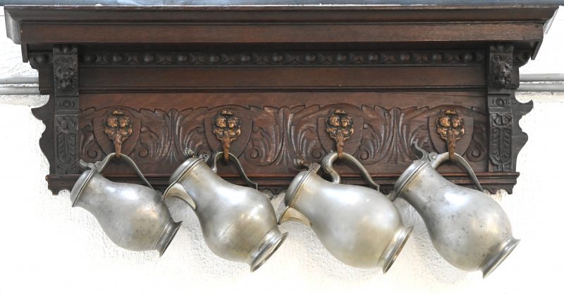 Een gebeeldhouwde eikenhouten kapstok versierd met leeuwenkoppen, met vier koperen haakjes, waaraan vier diverse beursvormige tinnen klepkannen. Omstreek 1900.