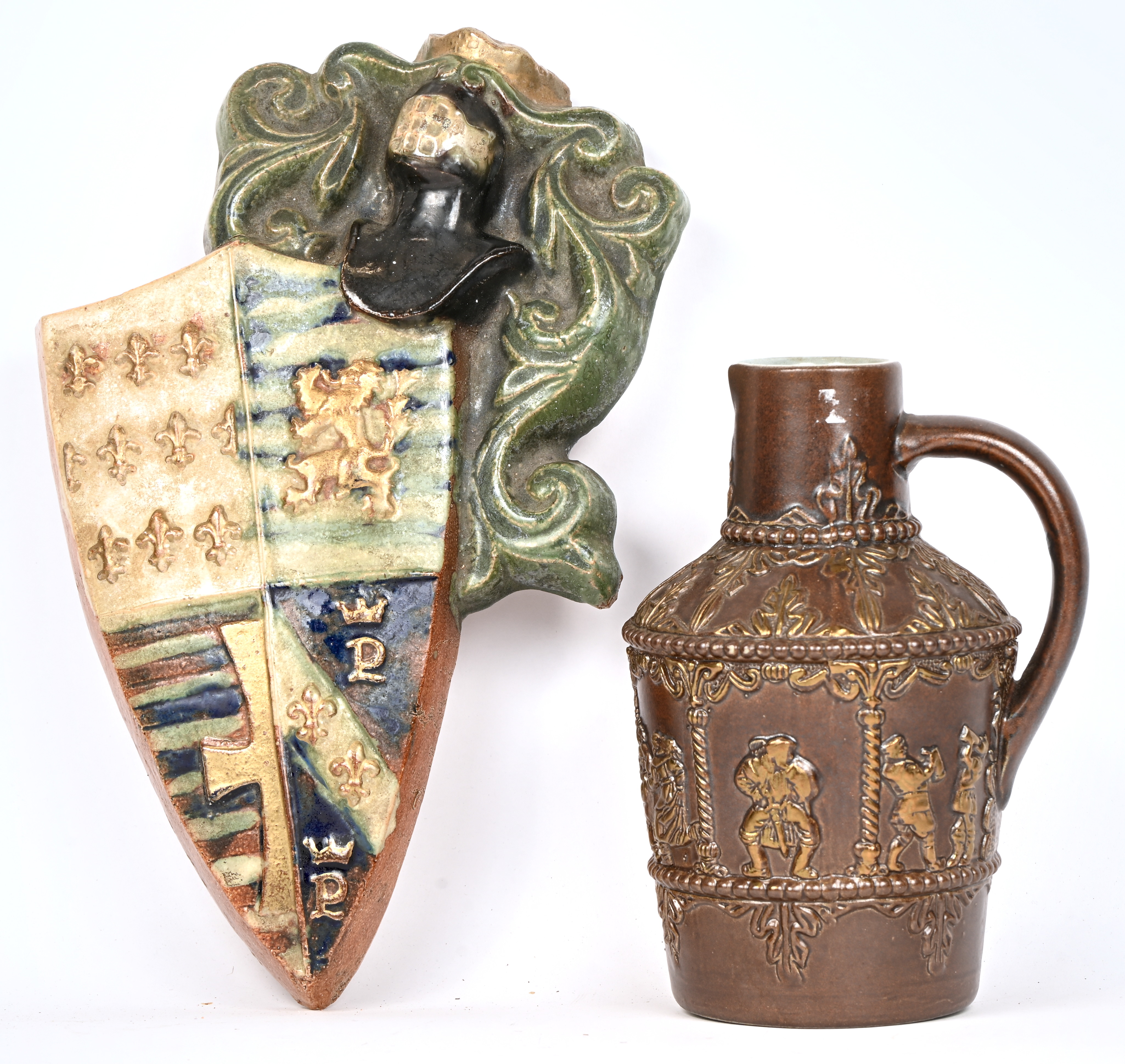 Een aardewerken wapenschild en een schenkkan versierd met middeleeuwse personages Een aardewerken wapenschild en een schenkkan versierd met middeleeuwse personages.