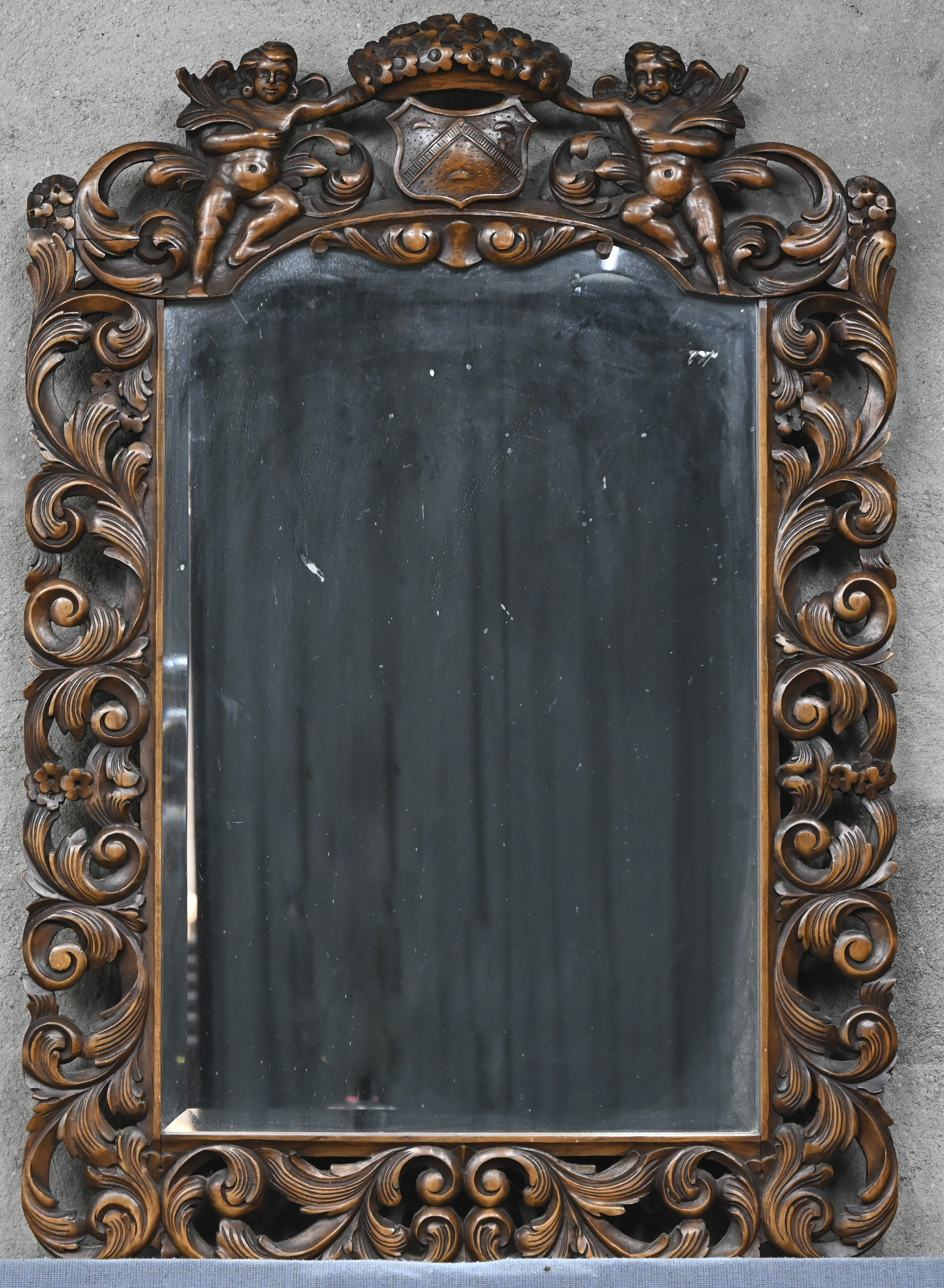 Een spiegel met gesculpteerd houten frame, neo-klassiek met wapenschild en twee cherubijnen Een spiegel met gesculpteerd houten frame, neo-klassiek met wapenschild en twee cherubijnen.
