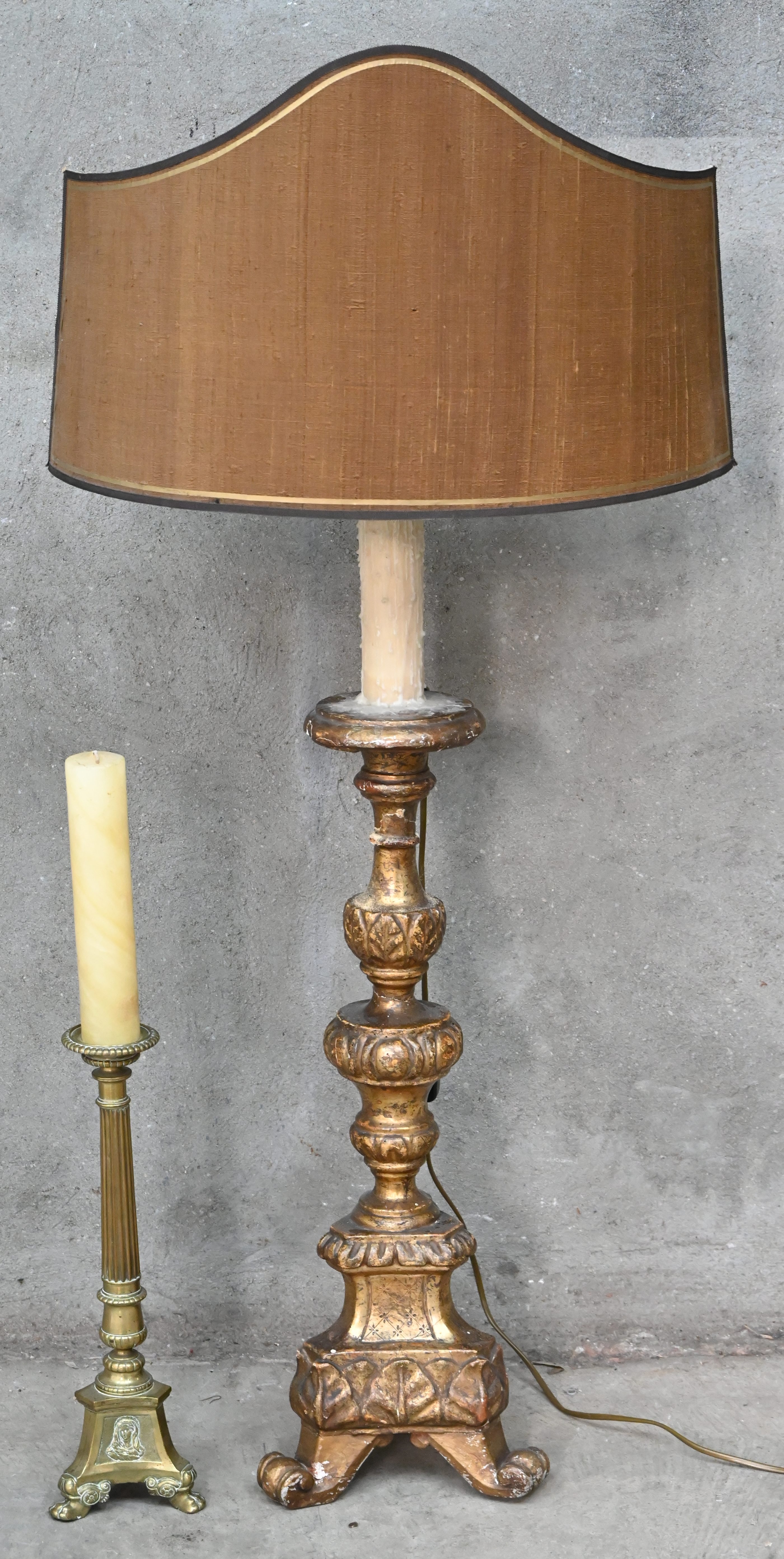 Een staande lamp in de vorm van een kandelaar van goudkleurig gepatineerd hout, met een halve lampenkap. Toegevoegd een messing kerkkandelaar Een staande lamp in de vorm van een kandelaar van goudkleurig gepatineerd hout, met een halve lampenkap. Toegevoegd een messing kerkkandelaar.