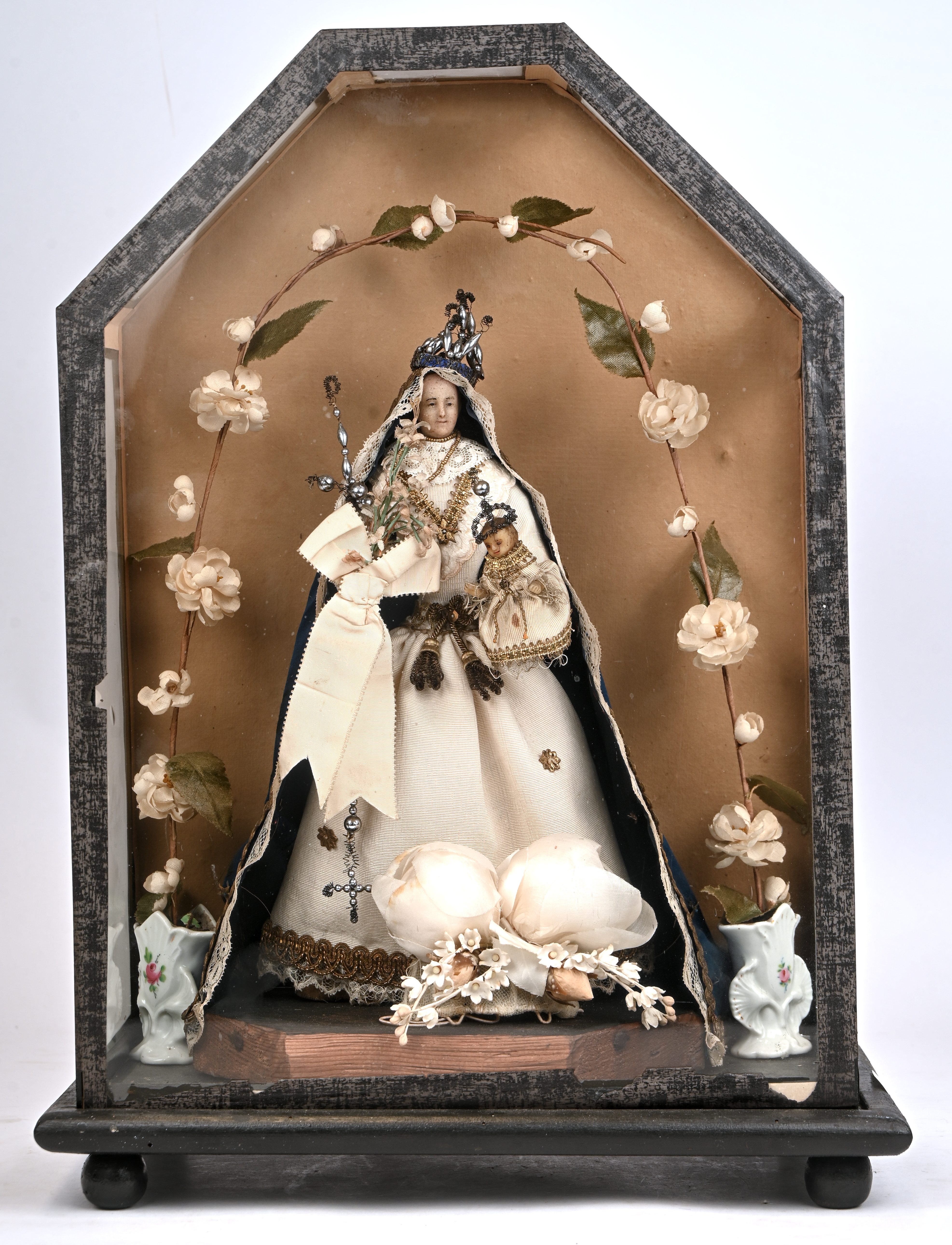 Een Madonna met wassen hoofd en met fijne gewaden onder presentatievitrinetje en omgeven door kunstbloemen. Volkskunst Een Madonna met wassen hoofd en met fijne gewaden onder presentatievitrinetje en omgeven door kunstbloemen. Volkskunst.