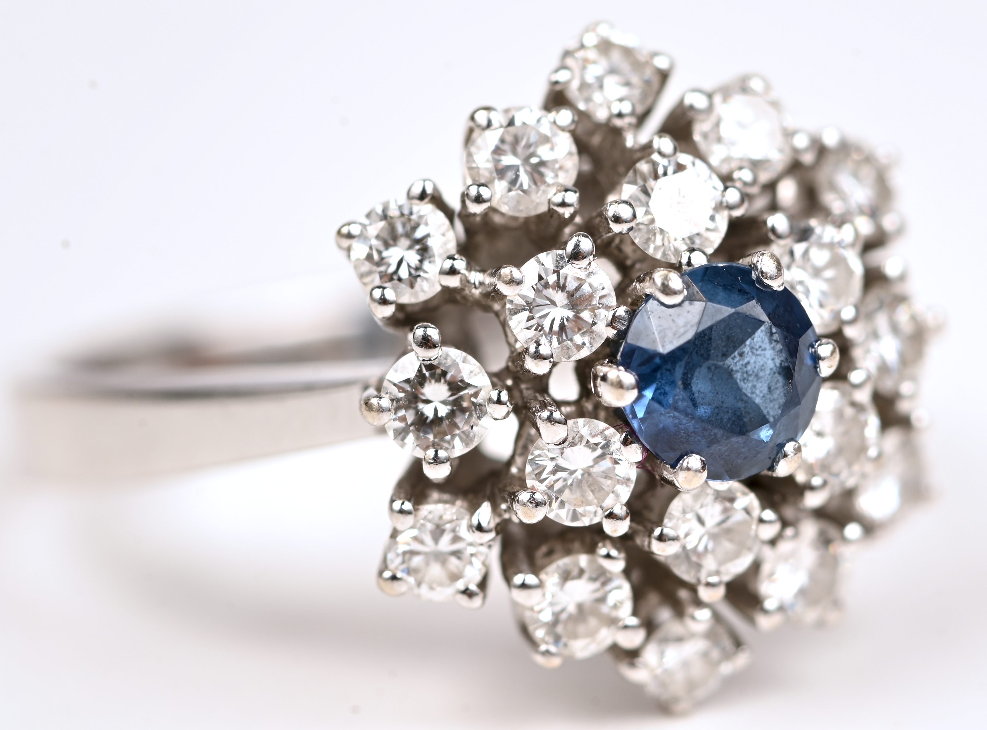 Een 14 kt witgouden ring bezet met een centrale saffier van +- 0,25 ct. en briljanten met een gezamenlijk gewicht van +- 1 ct Een 14 kt witgouden ring bezet met een centrale saffier van +- 0,25 ct. en briljanten met een gezamenlijk gewicht van +- 1 ct.
