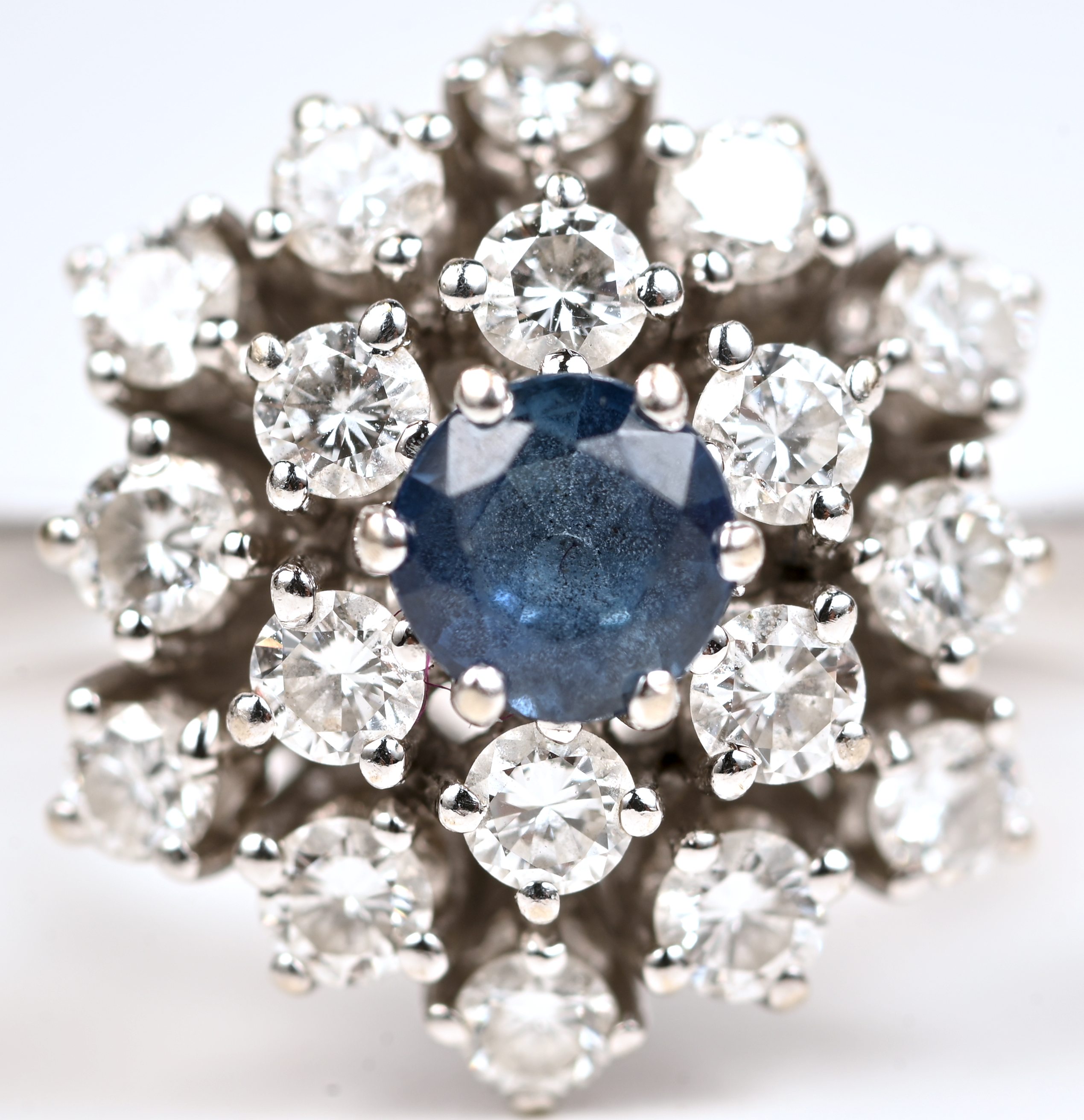 Een 14 kt witgouden ring bezet met een centrale saffier van +- 0,25 ct. en briljanten met een gezamenlijk gewicht van +- 1 ct Een 14 kt witgouden ring bezet met een centrale saffier van +- 0,25 ct. en briljanten met een gezamenlijk gewicht van +- 1 ct.
