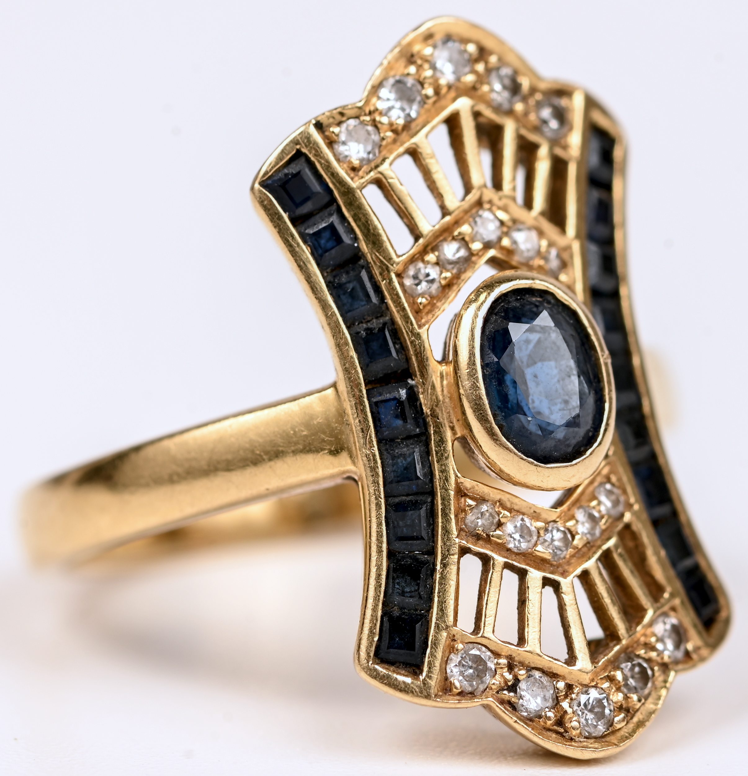 Een 18 kt art deco geelgouden ring bezet met een centrale saffier van +- 0,20 ct. en gekalibreerde saffieren met een gezamenlijk gewicht van +- 0,30 ct. en diamanten oude slijp met een gezamenlijk gewicht van +- 0,20 ct Een 18 kt art deco geelgouden ring bezet met een centrale saffier van +- 0,20 ct. en gekalibreerde saffieren met een gezamenlijk gewicht van +- 0,30 ct. en diamanten oude slijp met een gezamenlijk gewicht van +- 0,20 ct.