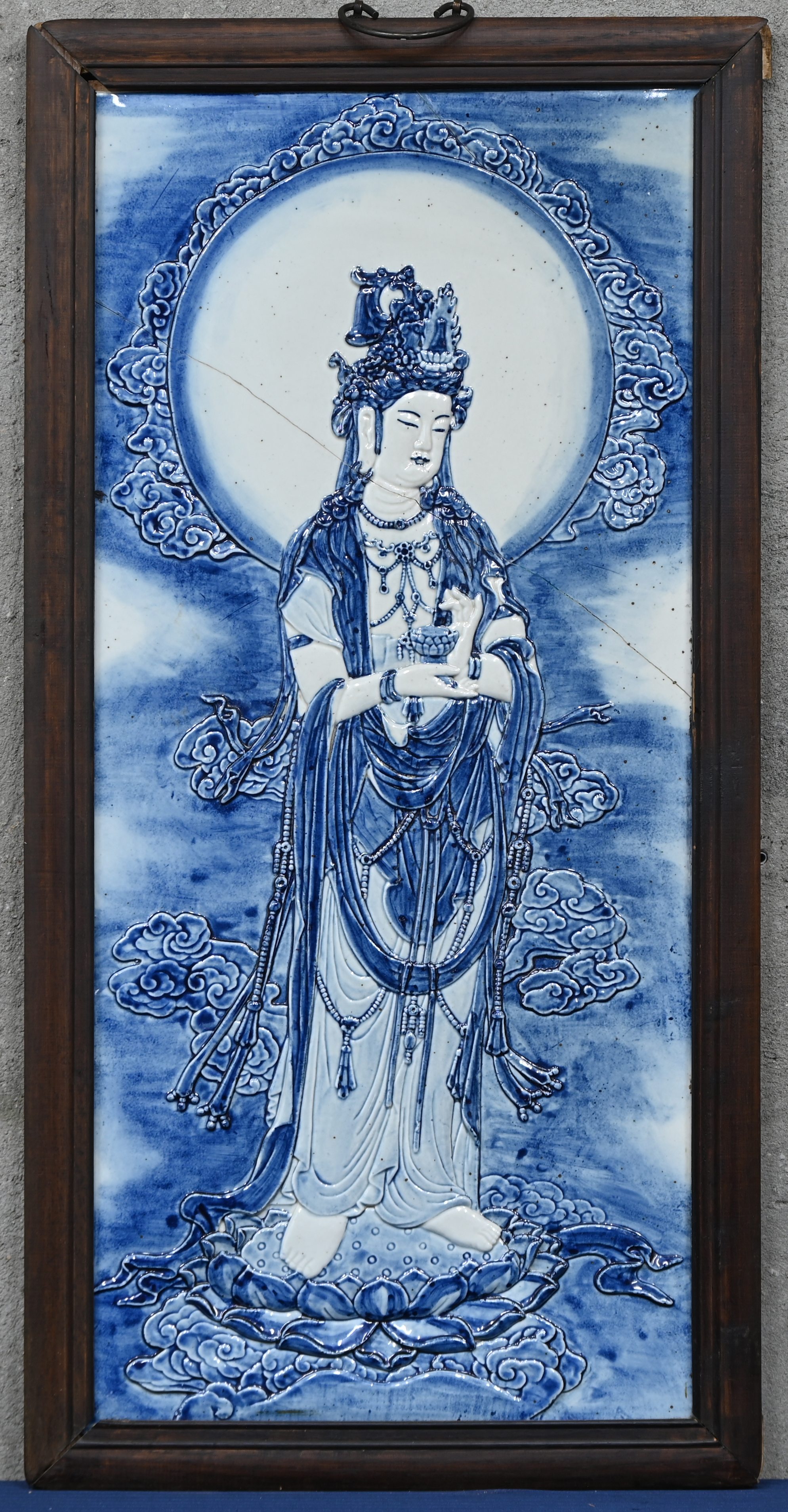 Een Chinese wandplaquette van blauw-wit porselein met een reliëfafbeelding van Guanyin in een houten lijst. De plaquette is doormidden gebroken Een Chinese wandplaquette van blauw-wit porselein met een reliëfafbeelding van Guanyin in een houten lijst. De plaquette is doormidden gebroken.