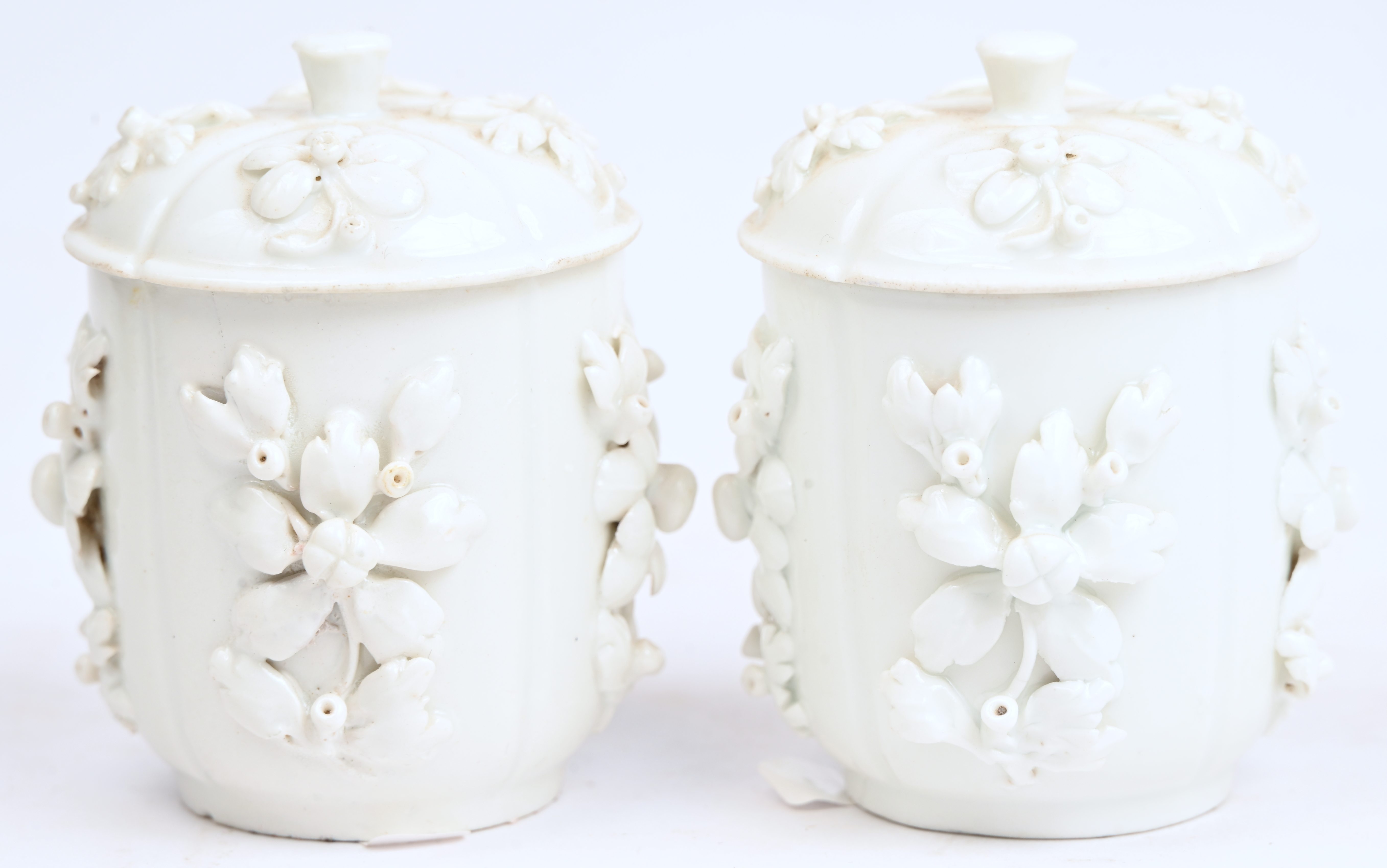 Een paar monochrome witte roompotjes met deksels,versierd met een bloemenmotief in reliëf. Onderaan gemerkt met de gekruiste zwaarden en kruisjes in goud Een paar monochrome witte roompotjes met deksels,versierd met een bloemenmotief in reliëf. Onderaan gemerkt met de gekruiste zwaarden en kruisjes in goud.